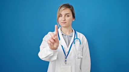 Young blonde woman doctor standing with serious expression saying no with finger over isolated blue background