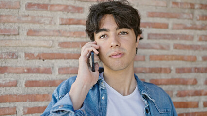 Young hispanic man talking on smartphone with serious expression over isolated brick background