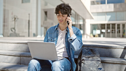 Young hispanic man student using laptop talking on smartphone at university