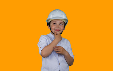 A child in a construction helmet with a pensive expression on an orange background. A cute boy in the role of a builder and a helmet looks thoughtfully to the side. Child profession concept.