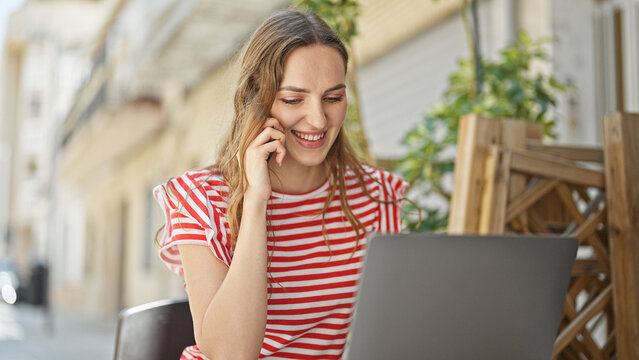 Young blonde woman talking on smartphone using laptop sitting on table at coffee shop terrace