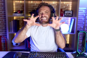 Middle age man with beard playing video games wearing headphones smiling funny doing claw gesture...