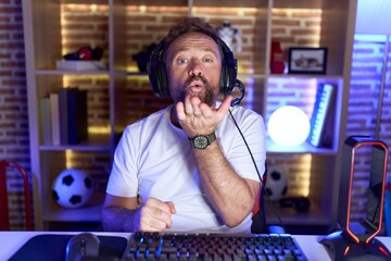 Middle age man with beard playing video games wearing headphones looking at the camera blowing a kiss with hand on air being lovely and sexy. love expression.