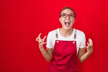 Young hispanic woman wearing waitress apron over red background crazy and mad shouting and yelling...