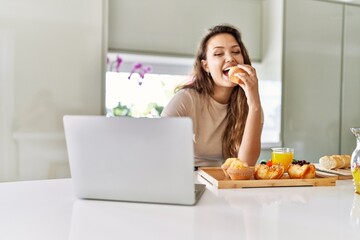 Young beautiful hispanic woman having breakfast using laptop at the kitchen