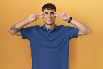 Young hispanic man standing over yellow background doing peace symbol with fingers over face, smiling cheerful showing victory