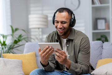 Young smiling hispanic man sitting on sofa at home wearing headphones and using tablet