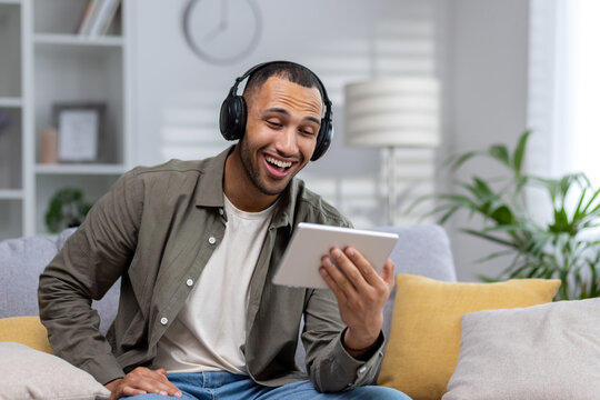 African american smiling young man sitting on couch at home wearing headphones and holding tablet, talking on phone call, watching video