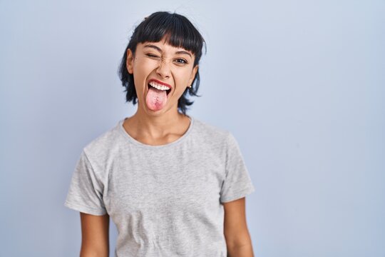 Young hispanic woman wearing casual t shirt over blue background sticking tongue out happy with funny expression. emotion concept.