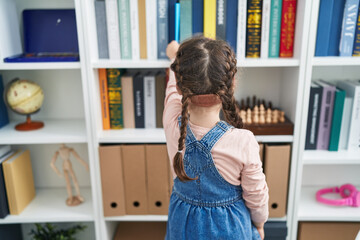 Adorable hispanic girl student looking for book standing backwards at classroom