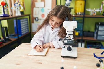 Adorable hispanic girl student using microscope writing notes at laboratory classroom