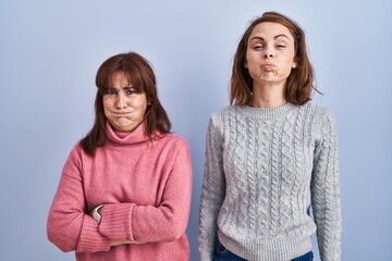 Mother and daughter standing over blue background puffing cheeks with funny face. mouth inflated with air, crazy expression.