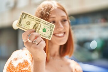 Young redhead woman smiling confident holding dollar at street