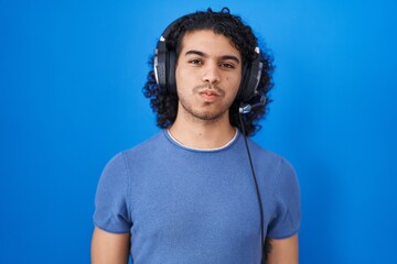 Hispanic man with curly hair listening to music using headphones puffing cheeks with funny face. mouth inflated with air, crazy expression. - obrazy, fototapety, plakaty