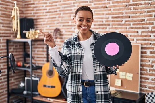 Beautiful african american woman holding vinyl record at music studio smiling happy pointing with hand and finger to the side