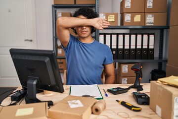 Hispanic man with curly hair working at small business ecommerce covering eyes with arm, looking...