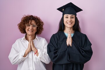 Hispanic mother and daughter wearing graduation cap and ceremony robe praying with hands together asking for forgiveness smiling confident.