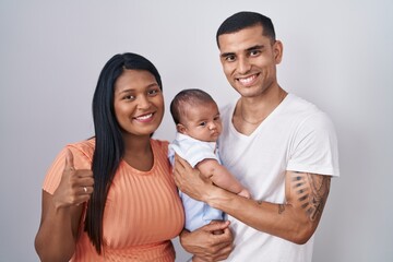 Young hispanic couple with baby standing together over isolated background doing happy thumbs up gesture with hand. approving expression looking at the camera showing success.