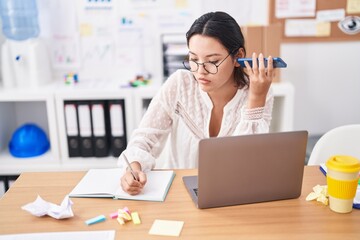 Young hispanic woman business worker listening voice message by smartphone writing on notebook at office