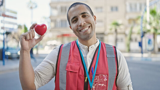 Young hispanic man volunteer smiling holding red heart for blood donation at street