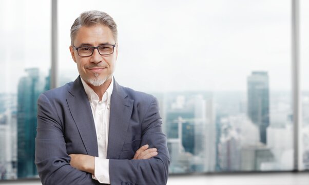 Business portrait - happy confident businessman in corporate skyscraper office, smiling. Mid adult, mature age man standing, looking at camera. Copy space.