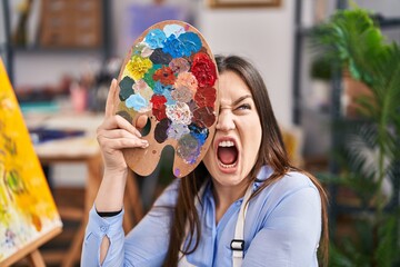Young brunette woman covering face with painter palette angry and mad screaming frustrated and furious, shouting with anger. rage and aggressive concept.