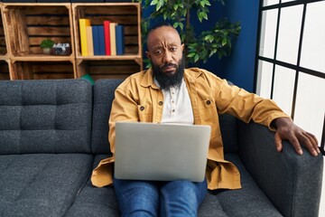 African american man using laptop at home sitting on the sofa thinking attitude and sober...