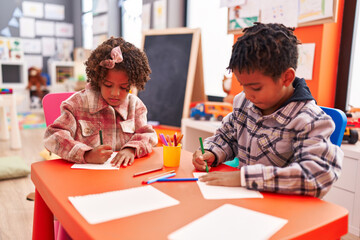 Adorable african american boy and girl preschool students sitting on table drawing on paper at...