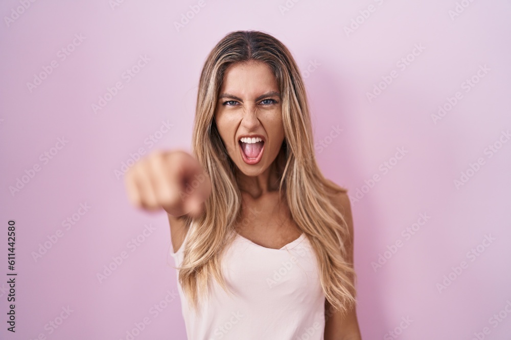 Wall mural Young blonde woman standing over pink background pointing displeased and frustrated to the camera, angry and furious with you