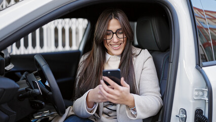 Young beautiful hispanic woman using smartphone sitting on car at street