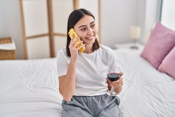 Fototapeta premium Young hispanic woman talking on the smartphone drinking wine at bedroom
