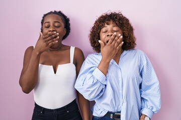 Two african women standing over pink background bored yawning tired covering mouth with hand. restless and sleepiness.