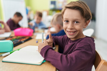 Adorable caucasian boy student writing on notebook doing thumb up gesture at classroom