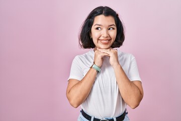 Young hispanic woman wearing casual white t shirt over pink background laughing nervous and excited with hands on chin looking to the side