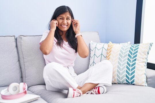 Young hispanic woman sitting on the sofa at home covering ears with fingers with annoyed expression for the noise of loud music. deaf concept.