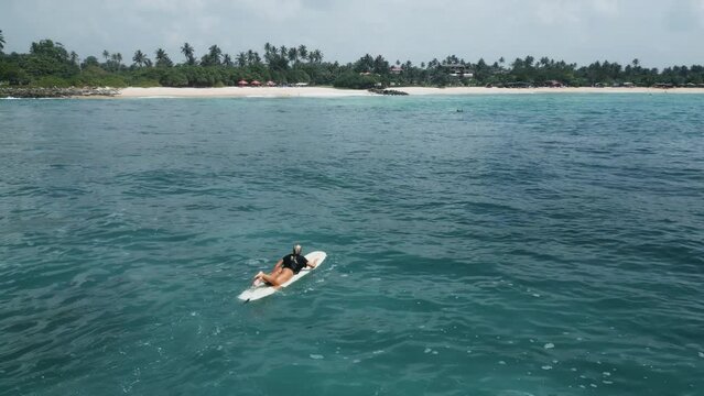 Young female surfer rowing on surfboard in crystal ocean water at scenic tropical beach on sunny day aerial view. Surfing girl enjoying sport paddling and surfing on summer vacation drone shot