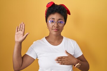 Young african american woman standing over yellow background swearing with hand on chest and open...