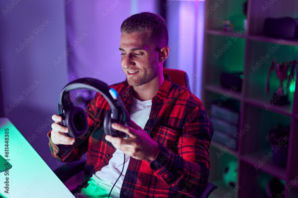 Poster young caucasian man streamer smiling confident holding headphones at gaming room