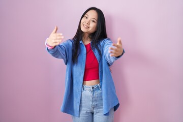 Young asian woman standing over pink background looking at the camera smiling with open arms for hug. cheerful expression embracing happiness.