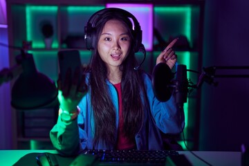 Young asian woman playing video games with smartphone with a big smile on face, pointing with hand finger to the side looking at the camera.