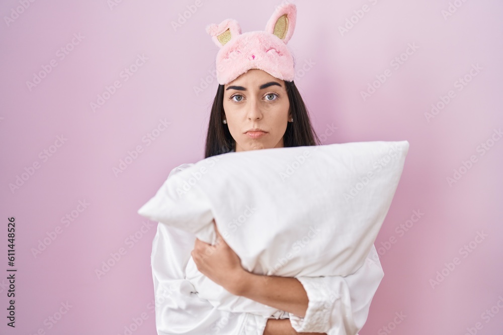 Poster young brunette woman wearing sleep mask and pajama hugging pillow depressed and worry for distress, 