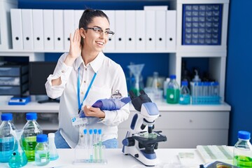 Young brunette woman working at scientist laboratory smiling with hand over ear listening an hearing to rumor or gossip. deafness concept.