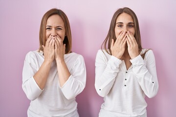 Middle age mother and young daughter standing over pink background laughing and embarrassed giggle covering mouth with hands, gossip and scandal concept