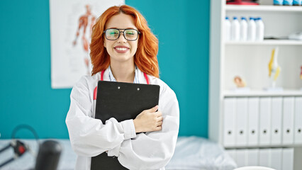 Young redhead woman doctor holding clipboard smiling at clinic