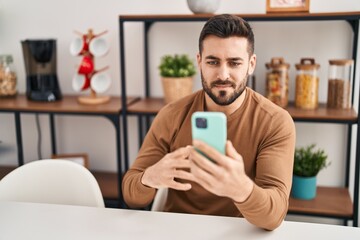 Young hispanic man using smartphone sitting on table at home