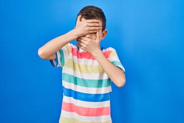 Young caucasian kid standing over blue background covering eyes and mouth with hands, surprised and shocked. hiding emotion