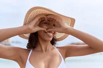 Young african american woman wearing summer hat doing heart gesture with hands at beach