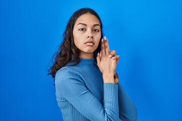 Young brazilian woman standing over blue isolated background holding symbolic gun with hand gesture, playing killing shooting weapons, angry face