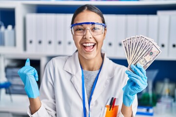 Young brazilian woman working at scientist laboratory holding money screaming proud, celebrating...