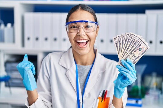 Young brazilian woman working at scientist laboratory holding money screaming proud, celebrating victory and success very excited with raised arms